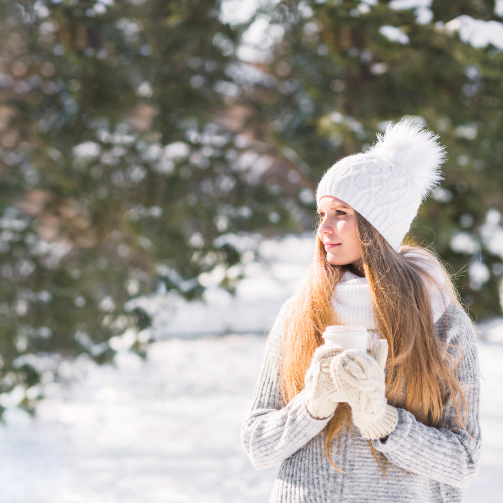 woman with long blond hair wearing a white bobble hat standing in snow holding a mug