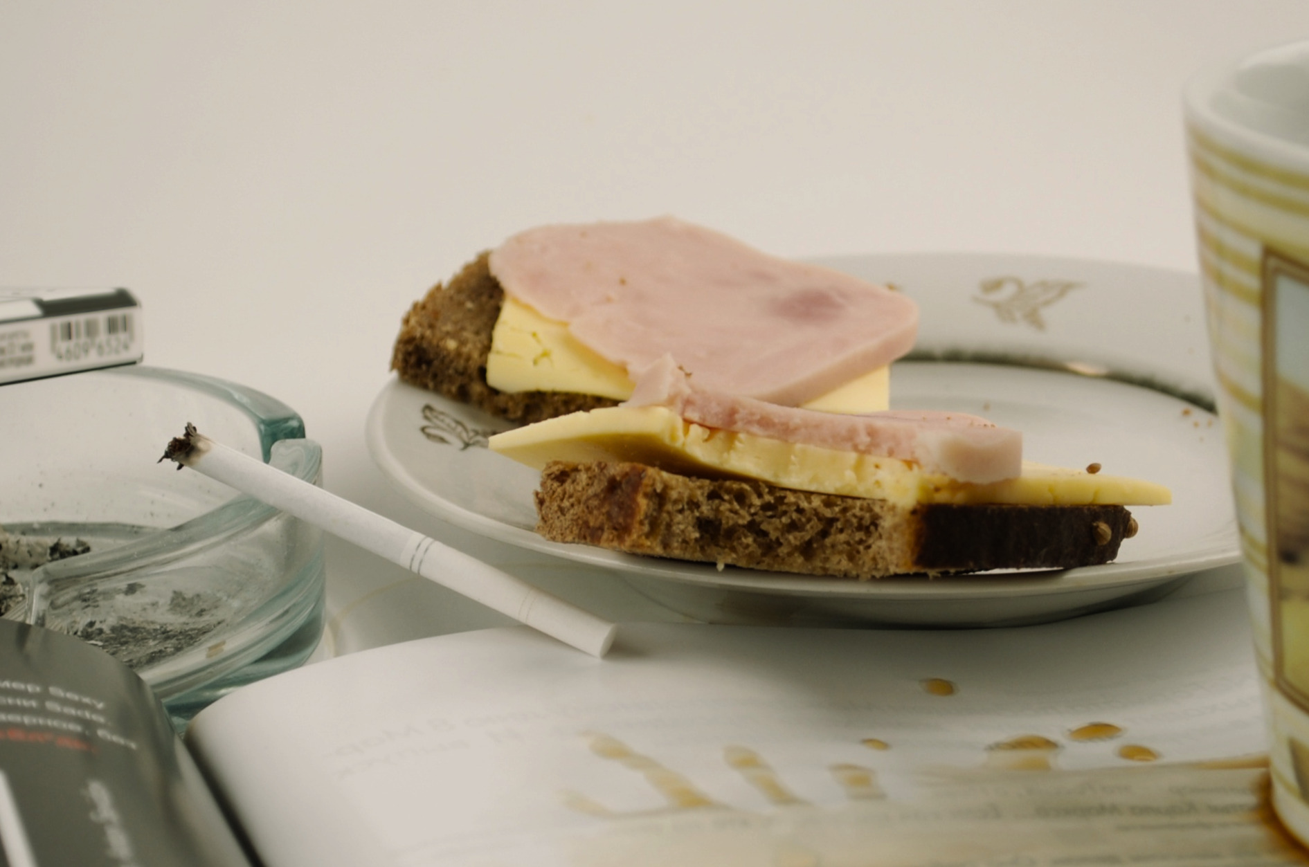 A slender cigarette propped on a glass ashtray next to open cheese and ham sandwiches